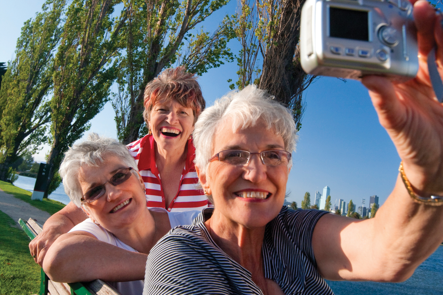 Three older women take a photo of themselves on a park bench.