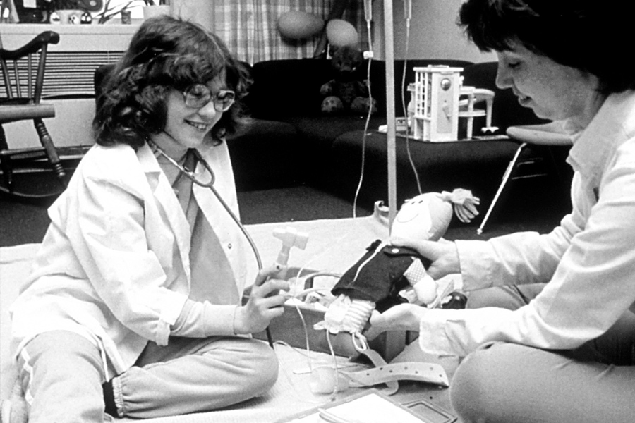 A female patient and nurse, both seated on the floor. The nurse is teaching the girl about procedures and techniques associated with chemotherapy.