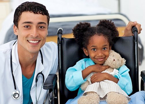 A doctor has his arm around a little girl sitting in a wheelchair and holding a stuffed bear. They are both smiling for the camera.
