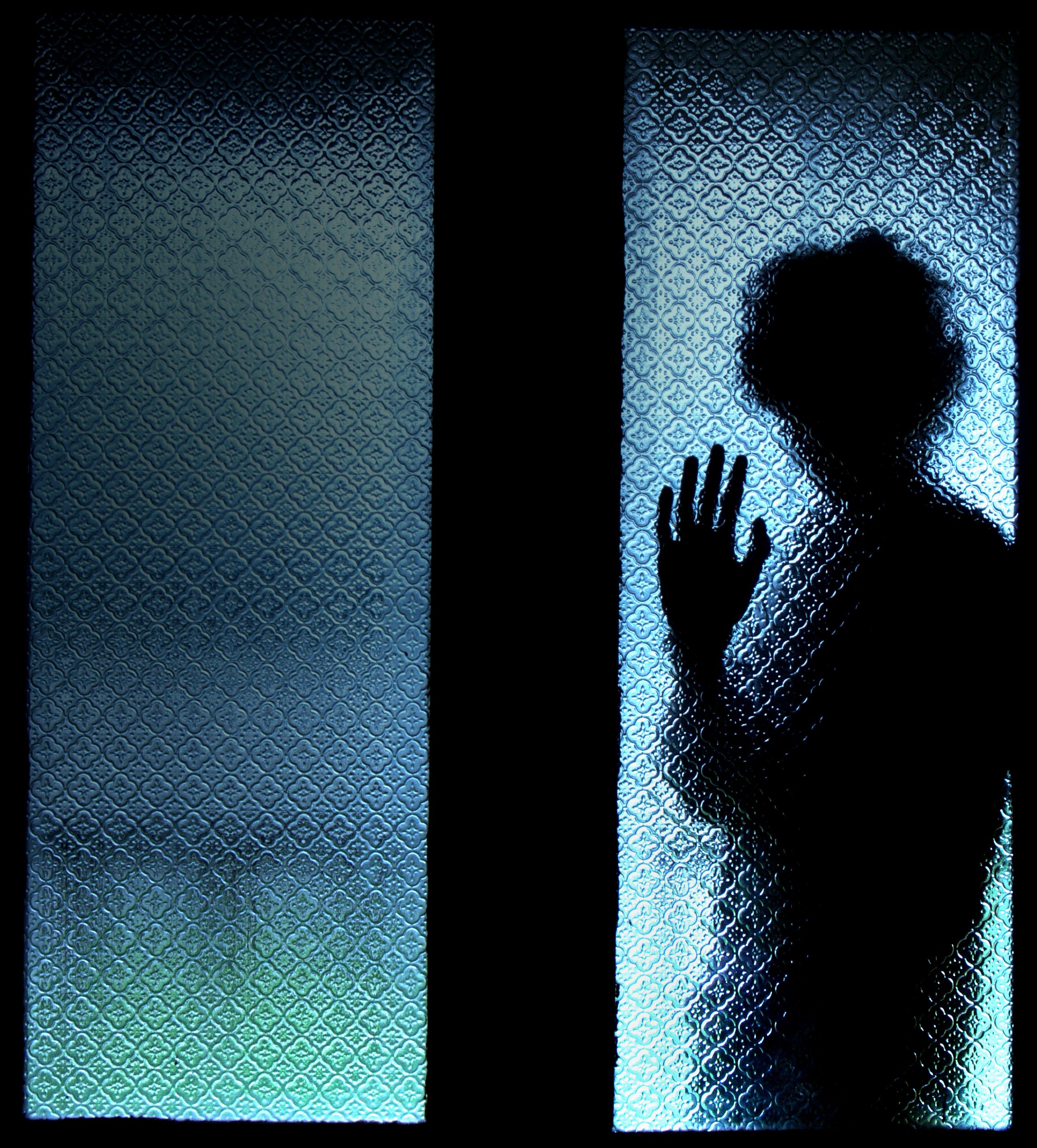 Silhouette of a man standing behind frosted glass holding up one hand while blue light shines behind him. 