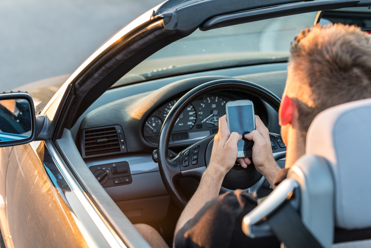 Boy using cell phone with two hands behind the driver’s wheel