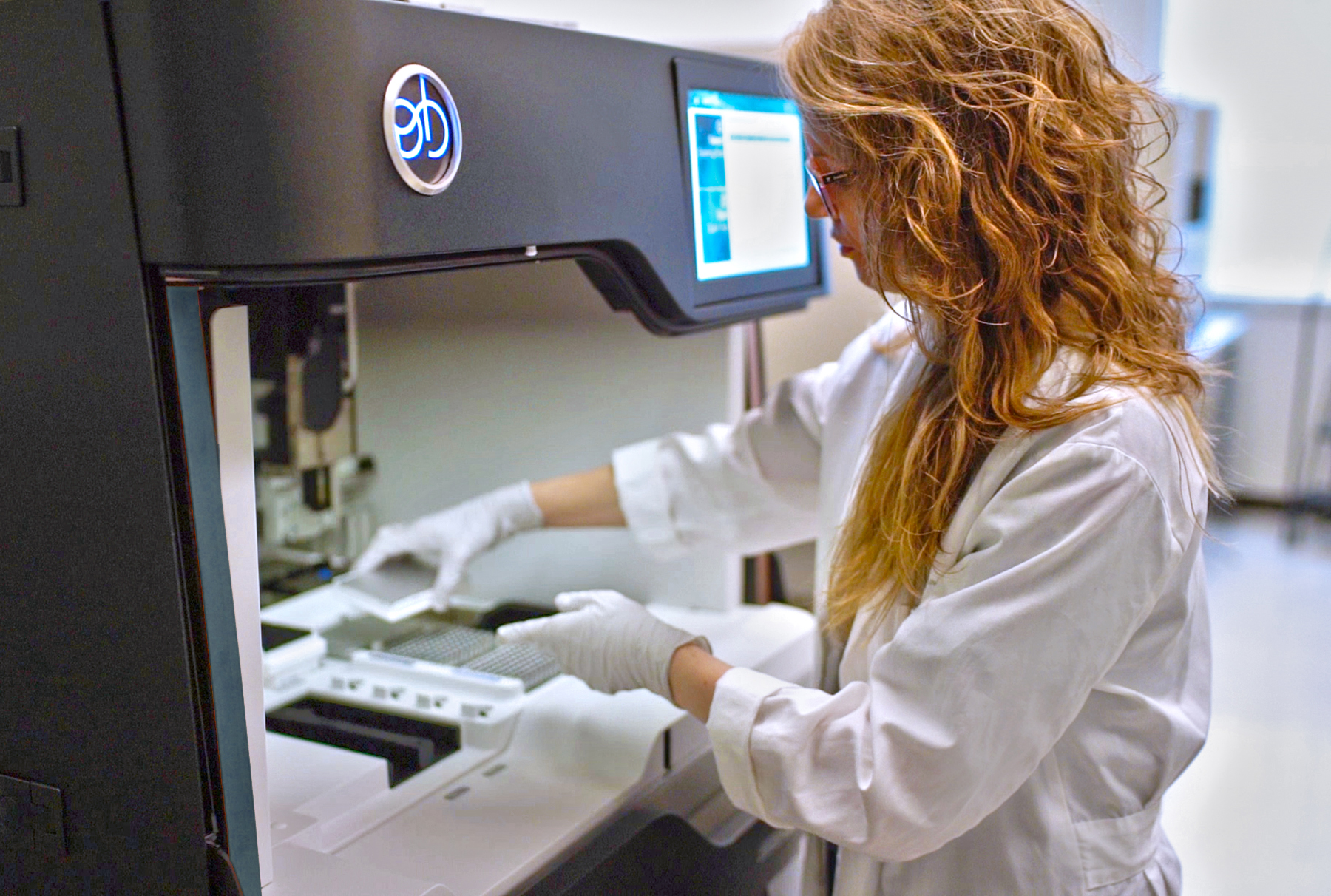 A young woman in a lab coat and gloves places a small tray of biosamples in a machine for DNA analysis.