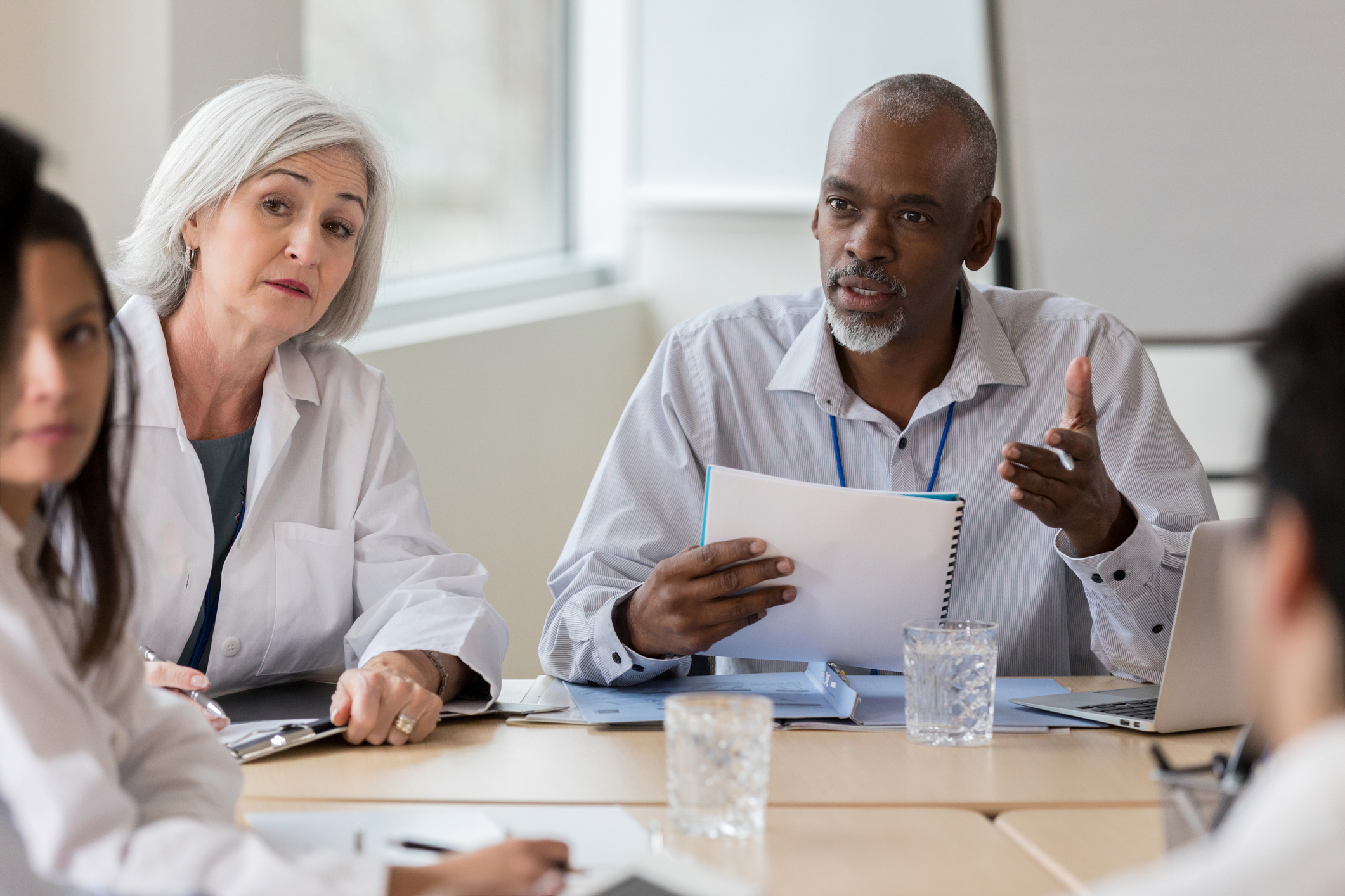 A senior businessman sits at a conference table during a medical staff meeting. He gestures as he speaks seriously with an unrecognizable coworker.