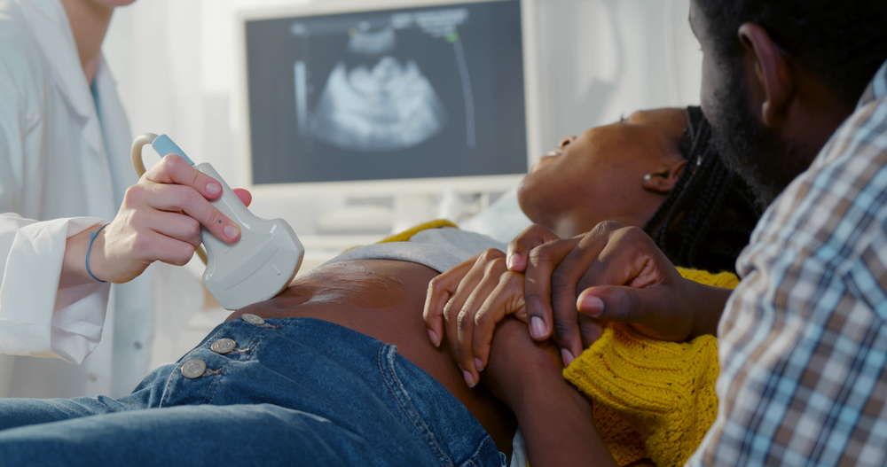 Afro pregnant couple smiling looking at sonogram results on monitor while on checkup at gynecologist. African husband holding hand of pregnant wife during ultrasound