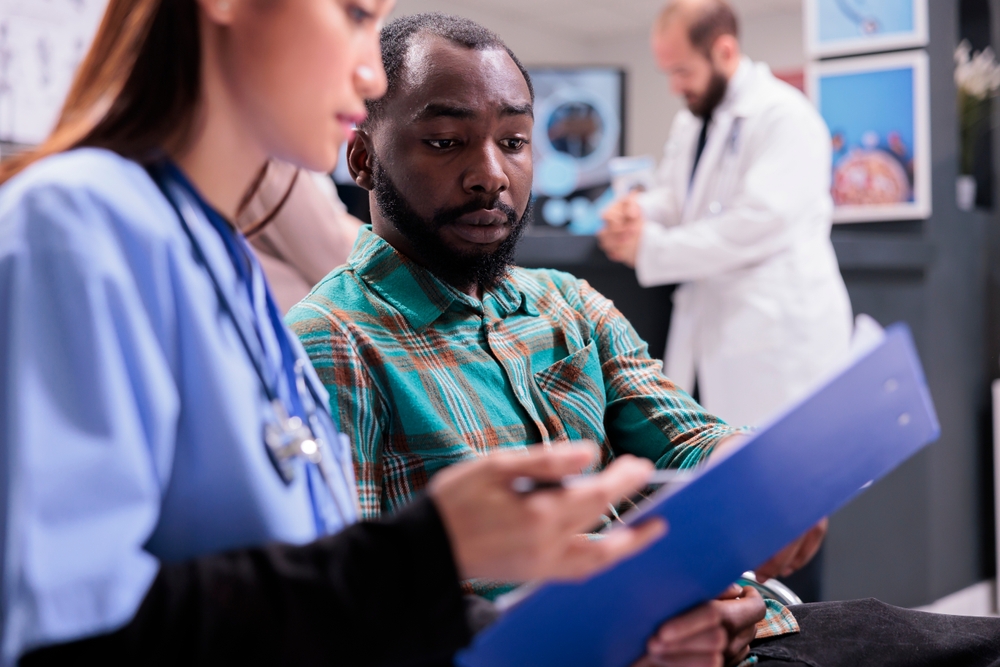 Close up of african american man reviewing medical records along with asian female member of in house hospital staff. Doctor indicating treatment to young male patient at sanatorium lobby. Close up of african american man reviewing medical records along with asian female member of in house hospital staff. Doctor indicating treatment to young male patient at sanatorium lobby.