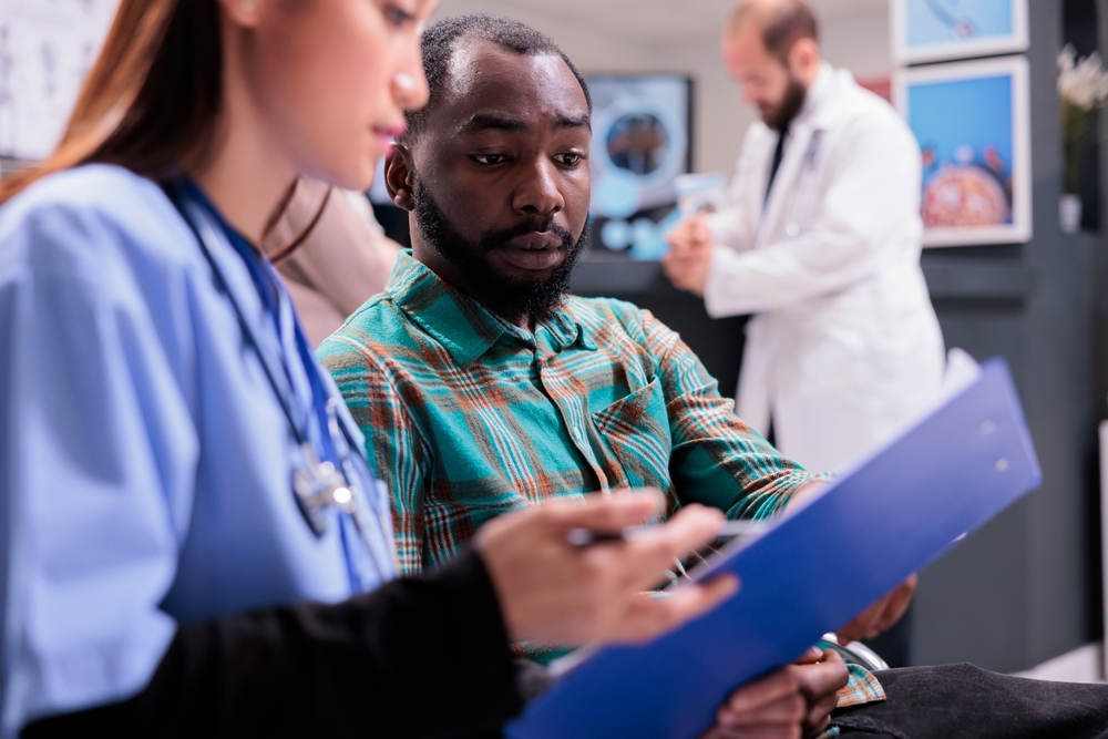 Close up of african american man reviewing medical records along with asian female member of in house hospital staff. Doctor indicating treatment to young male patient at sanatorium lobby.