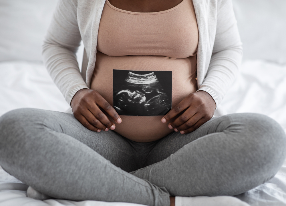 Unrecognizable black pregnant lady demonstrating her baby sonography photo while sitting on bed at home, showing firts photo of her child, enjoying happy maternity time, cropped image, closeup Unrecognizable black pregnant lady demonstrating her baby sonography photo while sitting on bed at home, showing firts photo of her child, enjoying happy maternity time, cropped image, closeup