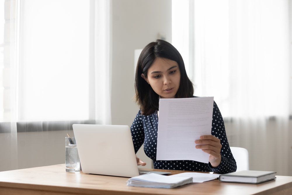 Attentive millennial asian female holding paper letter document reading financial statement. Focused vietnamese business woman worker employee review offer proposal study job contract terms conditions Attentive millennial asian female holding paper letter document reading financial statement. Focused vietnamese business woman worker employee review offer proposal study job contract terms conditions