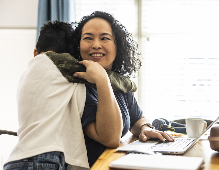 Woman sitting at home at a table working on her laptop and smiling while her pre-teen son hugs her.