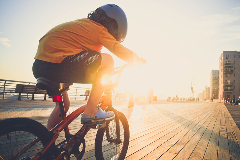 Young boy riding his bicycle on the boardwalk A young boy riding his bicycle on the boardwalk by the beach backlit by the sun wearing a helmet.