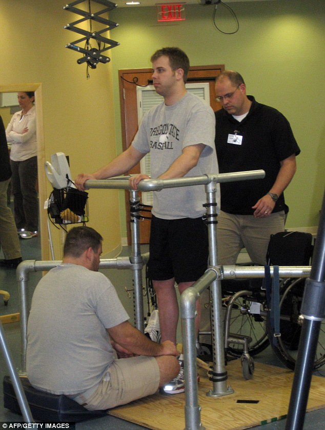 Photo of a standing man supported by hand rails and physical therapists Photo of a standing man supported by hand rails and physical therapists
