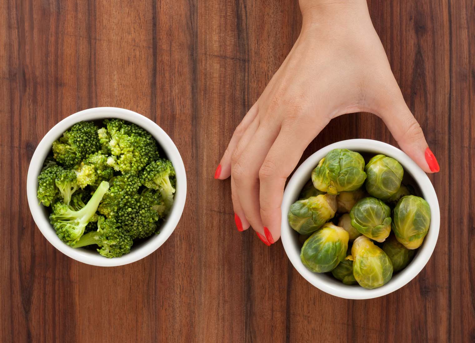 A woman’s hand holding a bowl of brussel sprouts, with a bowl of cooked broccoli to the left. A woman’s hand holding a bowl of brussel sprouts, with a bowl of cooked broccoli to the left.