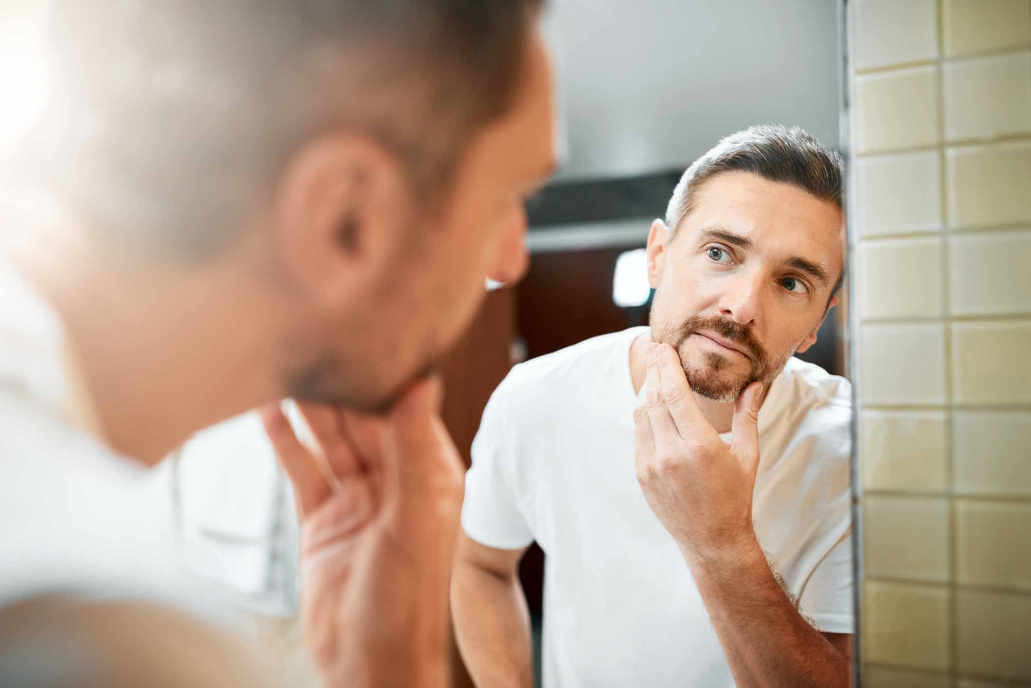 Mature man looking in bathroom mirror at the gray hair in his beard