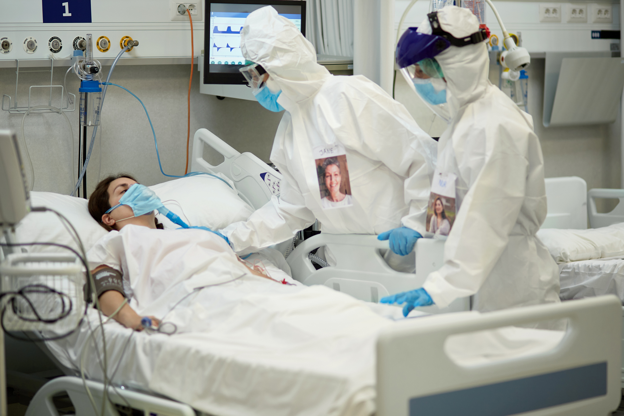 Medical team in protective equipment checking on female patient who is lying in a hospital bed wearing a surgical mask over a breathing device