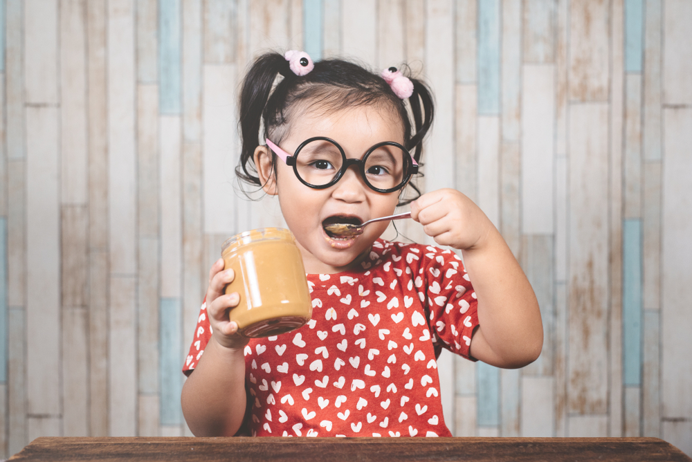 Young Asian girl holding a jar of peanut butter and putting a spoon in her mouth