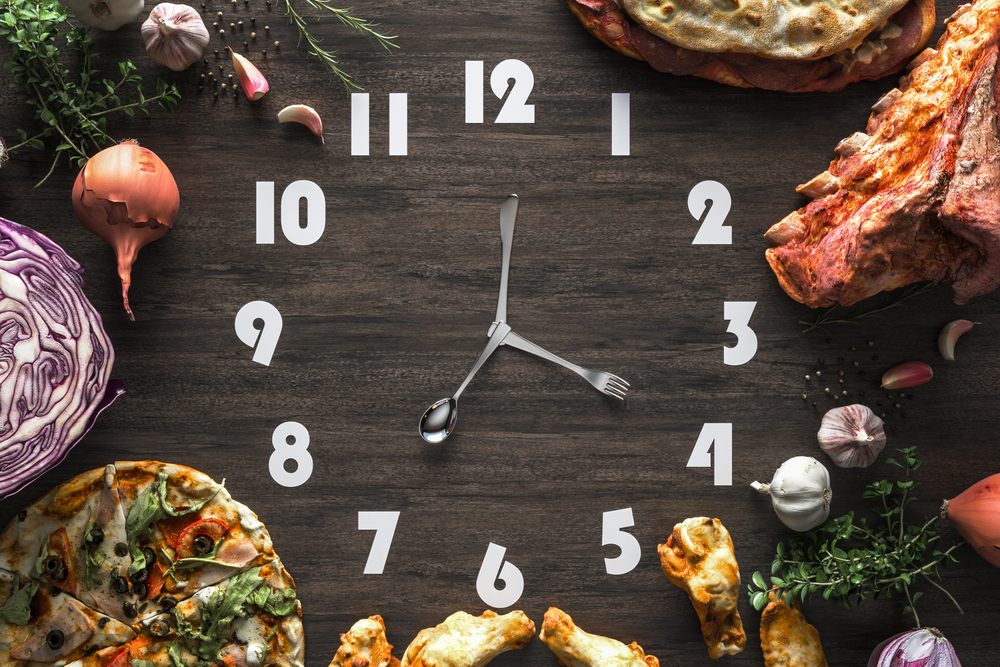Black wooden clock with utensils as hands and surrounded by food