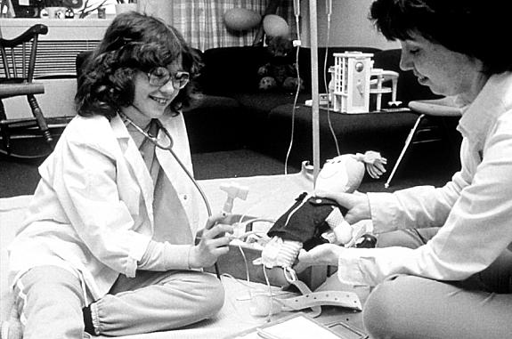 A female patient and nurse, both seated on the floor. The nurse is teaching the girl about procedures and techniques associated with chemotherapy.