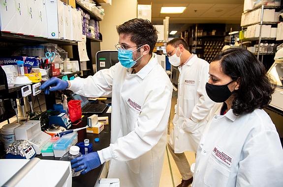 Three scientists wearing lab coats are standing near a lab bench and looking at a tool. 