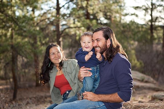 An American Indian couple hold their smiling baby outside, surrounded by trees.