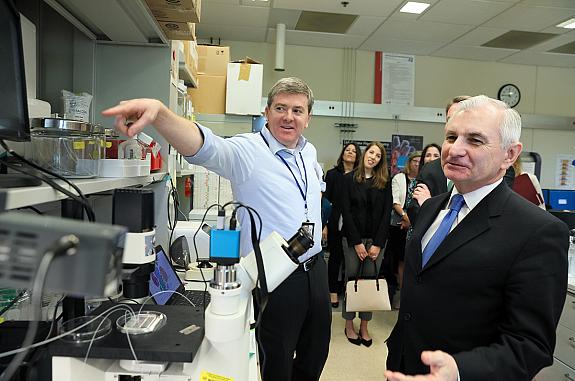 Two men, scientist and Senator, look at a scientific device designed to perform single cell analysis while staff look on.