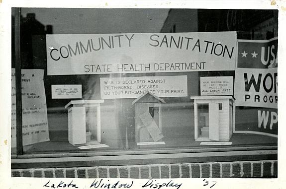 Exterior view of window display showing three model privies under a sign that reads "Community Sanitation."