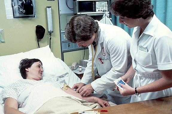 An adult woman lying in a hospital bed, covered with a sheet. She is having blood drawn by a male doctor with a female nurse in attendance. The patient is looking at the doctor and smiling.