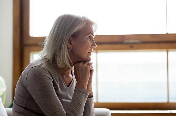 Side view of thoughtful middle-aged mature woman sitting on a couch at home in front of a window.