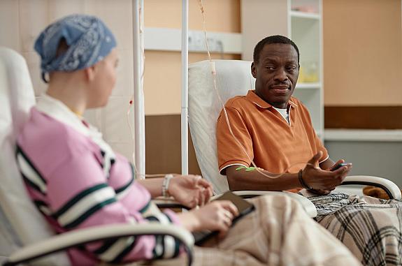 A black man and a white woman with a head scarf talking during chemotherapy treatment in clinic.