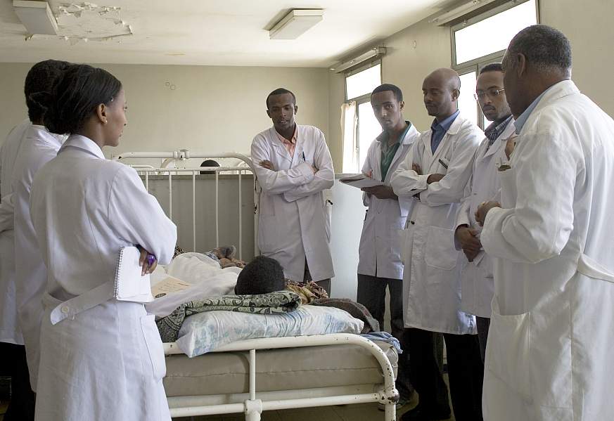 Medical students in white coats gather and discuss around patient bed in hospital