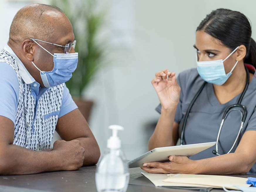 A healthcare worker wearing a medical face mask speaking to a masked patient and referencing a tablet.