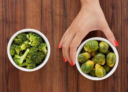 A woman’s hand holding a bowl of brussel sprouts, with a bowl of cooked broccoli to the left.