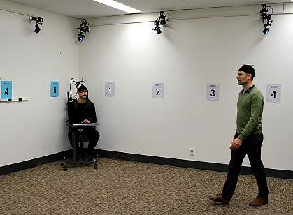 Picture of volunteer epilepsy patient sitting in corner of experimental room, wearing a special brain wave monitoring backpack and watching another participant search for a hidden spot. The other participant is shown walking.