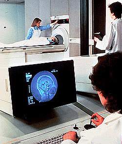 Image of brain on computer screen in foreground, with patient in background lying on an MRI machine
