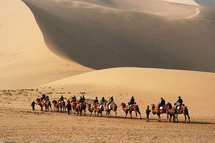 Photo of people riding camels past sand dunes