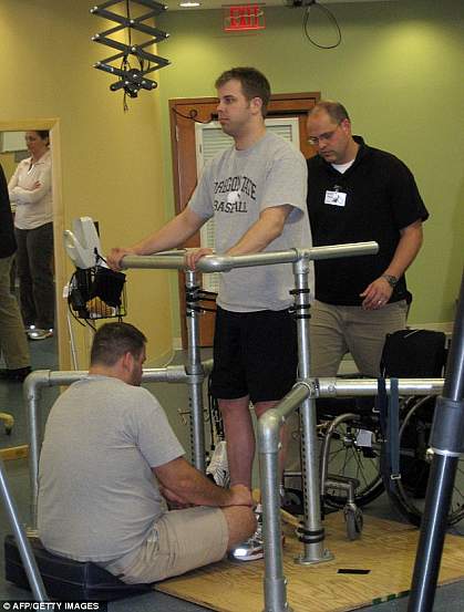 Photo of a standing man supported by hand rails and physical therapists