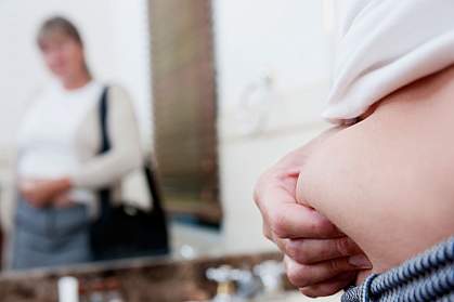 Woman looking at her reflection in a bathroom mirror and pinching her stomach fat.