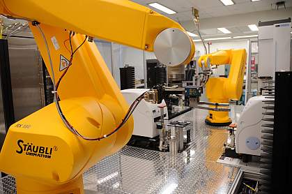 A robot arm (foreground) retrieves assay plates from incubators to place them at compound transfer stations or hand them off to another arm (background).