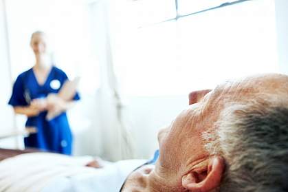 Elderly man in hospital bed with a blurred image of a nurse in the background