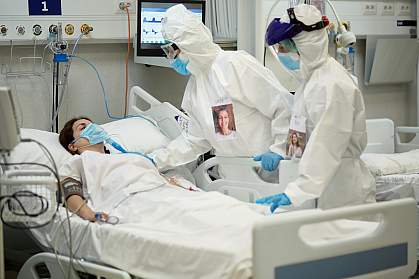 Medical team in protective equipment checking on female patient who is lying in a hospital bed wearing a surgical mask over a breathing device