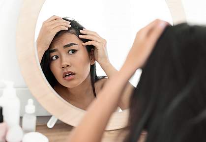 Troubled young Asian woman checking for thinning hair in mirror