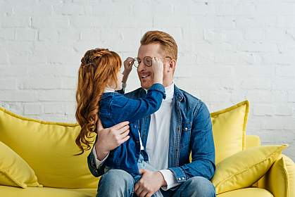 Red-haired daughter and father playing with his glasses while sitting on sofa