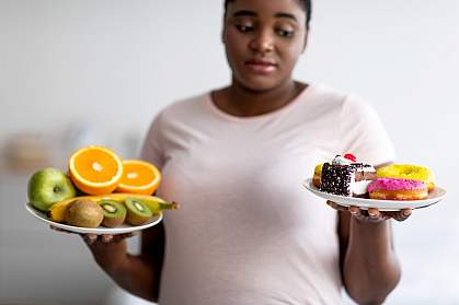 Black woman holding plates with one plate with fruits and one with cakes and donuts, looking at the unhealthy plate with skepticism.