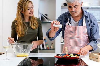 Man adding a pinch of salt to a pan of vegetables while woman whisks eggs.