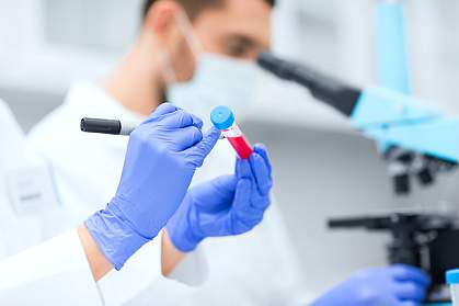 Laboratory scene with scientists’ hands labelling a test tube while another researcher in the background looks through a microscope.