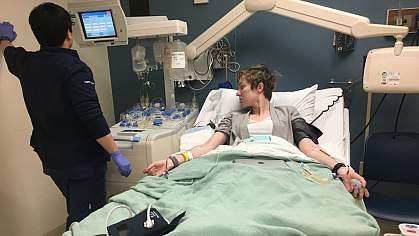 A medical technician watches the monitor of a woman with IVs in a hospital bed.