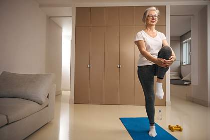 Older woman standing on one leg on a mat in the living room.