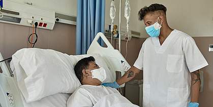 Young male nurse in white scrubs standing at bedside with hand on shoulder of recovering mature male COVID-19 patient, both wearing protective masks.