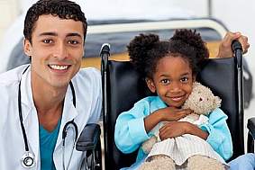 A doctor has his arm around a little girl sitting in a wheelchair and holding a stuffed bear. They are both smiling for the camera.