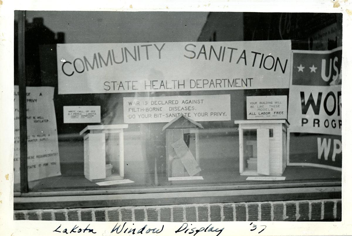 Exterior view of window display showing three model privies under a sign that reads “Community Sanitation”