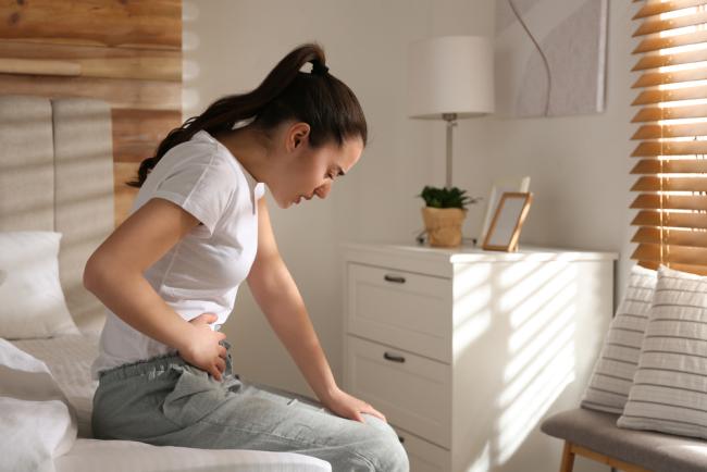 Young woman sitting on bed and clutching her stomach in pain.